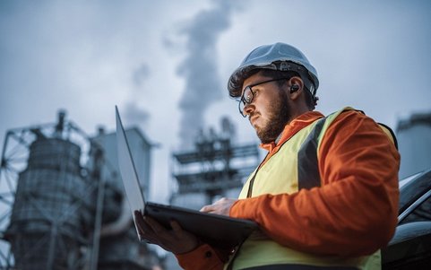An employee wearing a high-visibility jacket and white helmet, looking at a laptop in front of a factory.