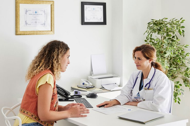 Ärztin im weißen Kittel im Gespräch mit einer Patientin an einem Schreibtisch in einer hellen Praxis.