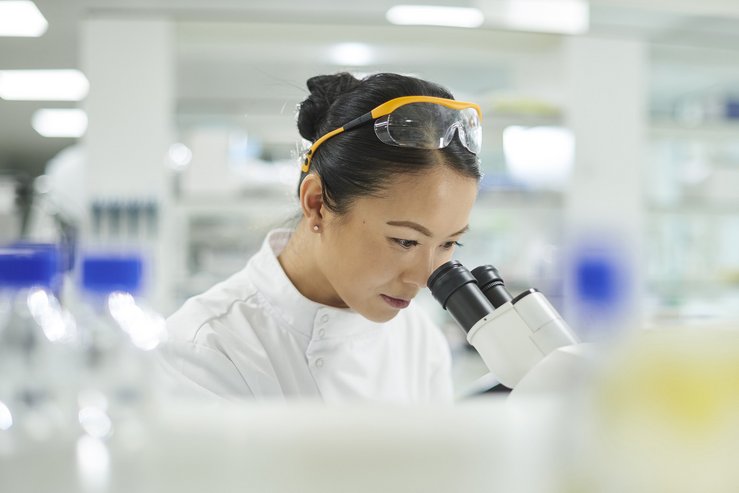 Lab technician wearing safety glasses conducts microscope analysis in a laboratory setting.