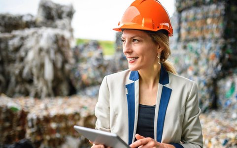 Frau mit orangefarbenem Schutzhelm und Blazer hält ein Tablet auf einem Recyclinghof.