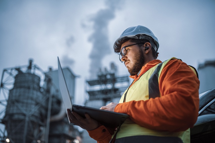 An employee wearing a high-visibility jacket and white helmet, looking at a laptop in front of a factory.